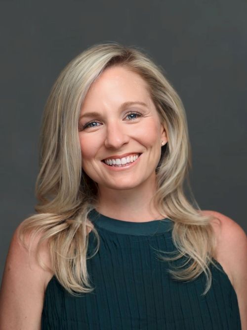 A person with blonde hair and a teal top is smiling against a neutral background.