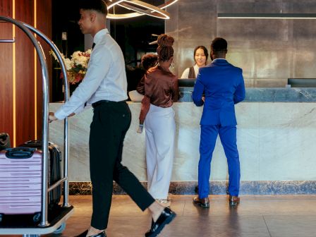 A reception area with two people checking in, a bellhop with luggage, and a modern light fixture overhead.