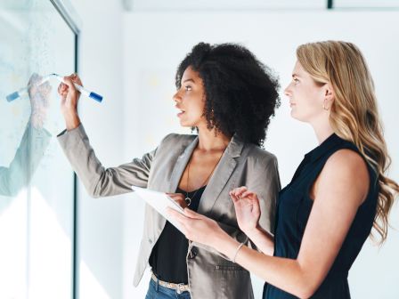 Two people are collaborating at a whiteboard, one writing with a marker while the other holds a notebook, both focused on a project.