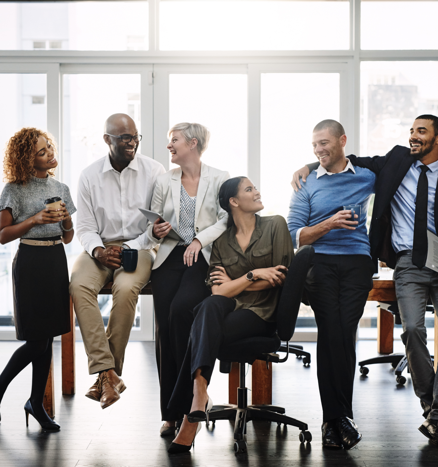 A group of six people in business attire are smiling and conversing in an office setting, holding coffee cups and appearing relaxed.
