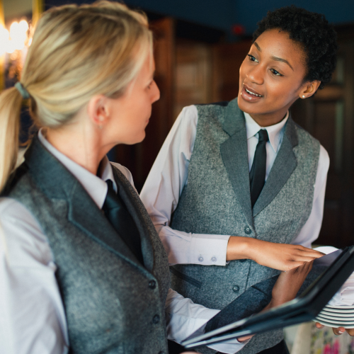 Two people in uniforms are having a discussion while one holds a notepad, seemingly in a restaurant or service setting.