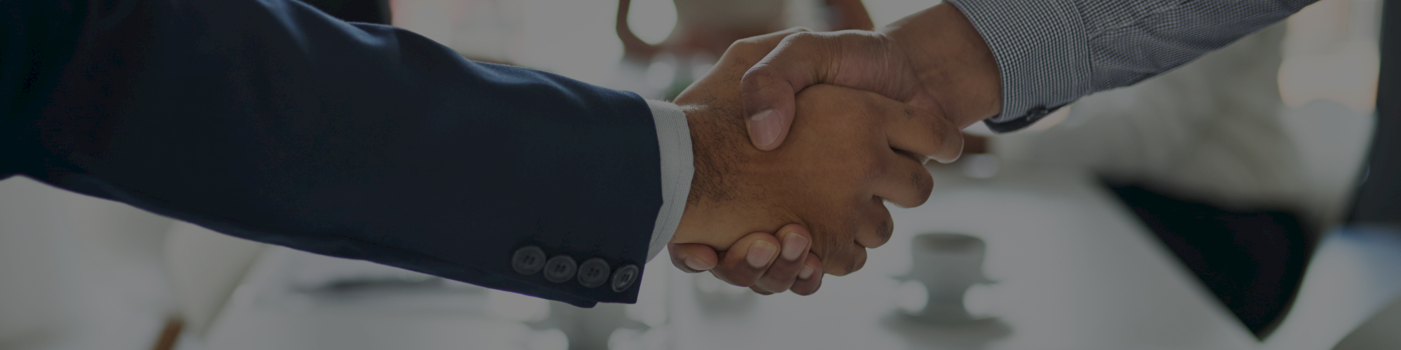 Two people are shaking hands in the foreground, with a blurred meeting scene in the background, suggesting a business setting.